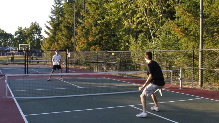 Player in a ready stance at the baseline preparing to return, with opponent and yellow pickleball across the net