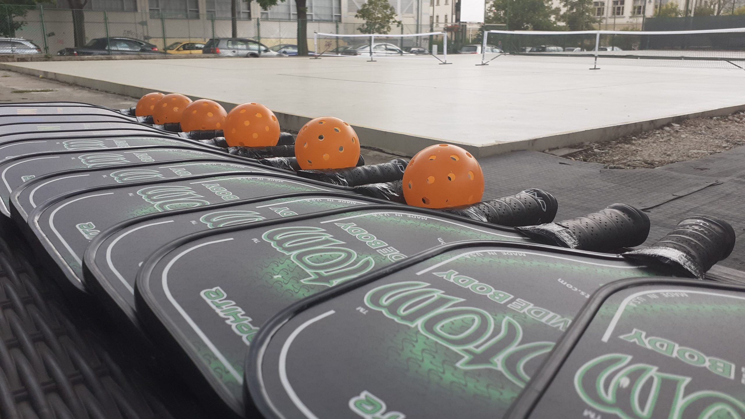 Row of graphite wide-body pickleball paddles and orange balls lined up courtside, with outdoor nets behind them
